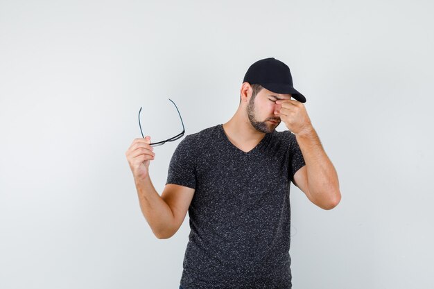 Young man taking off glasses, massaging eyes in t-shirt and cap and looking fatigued