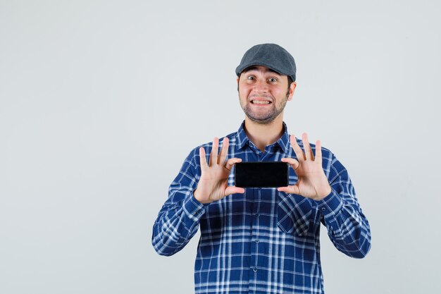 Молодой человек фотографирует на мобильном телефоне в рубашке, кепке и выглядит веселым, вид спереди.