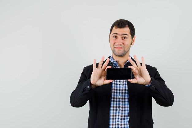 Young man taking photo on mobile phone in shirt, jacket and looking jolly , front view.