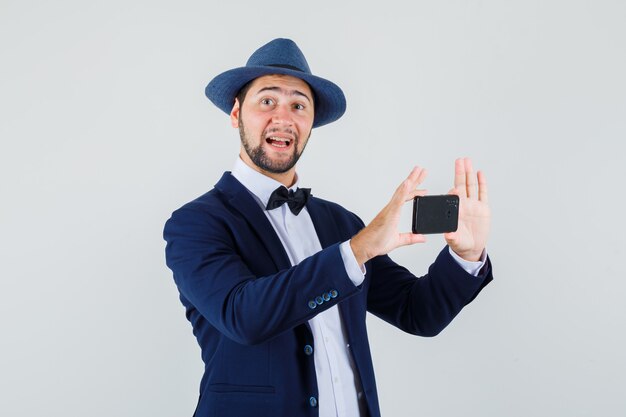 Young man taking photo on mobile phone in suit, hat and looking optimistic. front view.