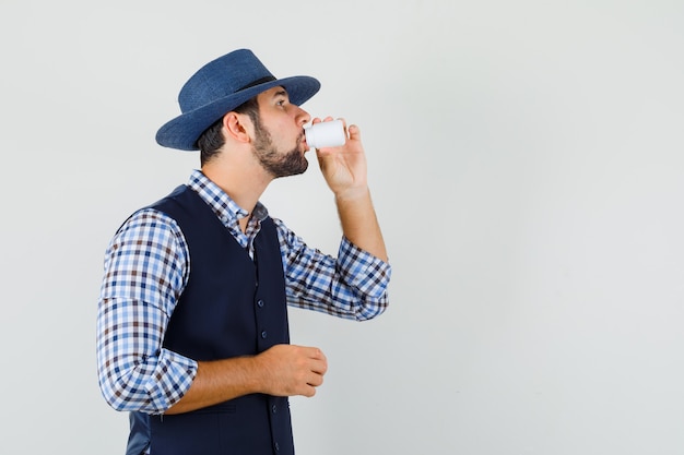 Young man taking pills in shirt, vest, hat