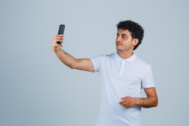 Молодой человек принимает селфи на мобильном телефоне в белой футболке и выглядит мило, вид спереди.