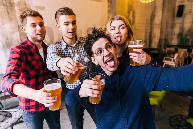 Young man taking selfie on mobile phone with his friends holding the glasses of beer
