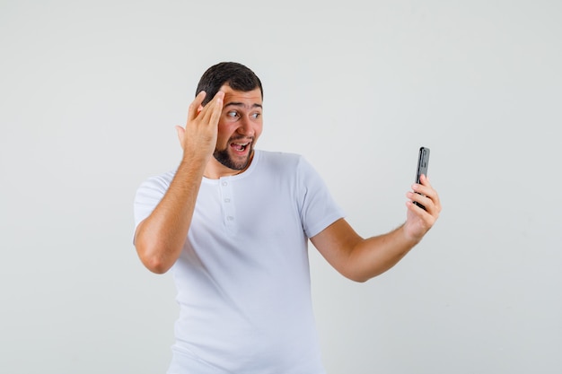 Young man taking selfie in white t-shirt and looking glad. front view.