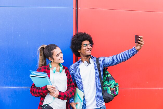 Young man taking selfie with his girlfriend holding books in hand on mobile phone against red and blue wall
