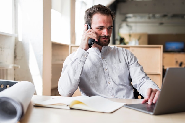 Free photo young man talking on cellphone while working on laptop