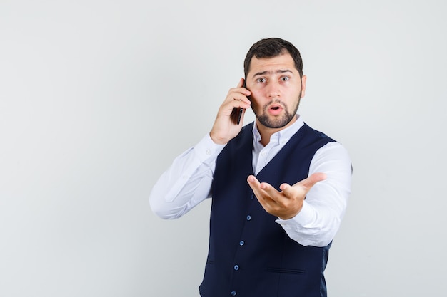 Young man talking on cellphone with raised hand in shirt and vest and looking puzzled