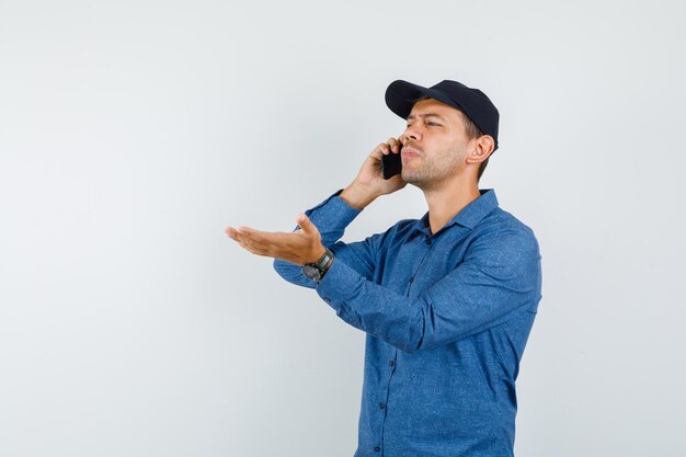 Молодой человек разговаривает по мобильному телефону в голубой рубашке, кепке и выглядит смущенным, вид спереди.