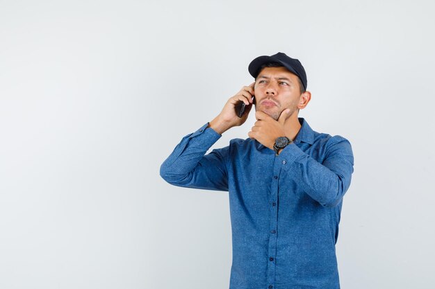 파란색 셔츠, 모자와 생각에 잠겨 찾고 휴대 전화에 얘기 하는 젊은 남자. 전면보기.