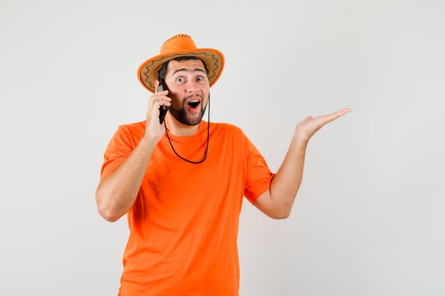 オレンジ色のTシャツ、帽子、幸せそうに見える携帯電話で話している若い男。正面図。