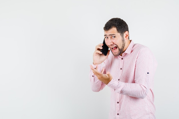 Молодой человек разговаривает по мобильному телефону в розовой рубашке и выглядит сердитым