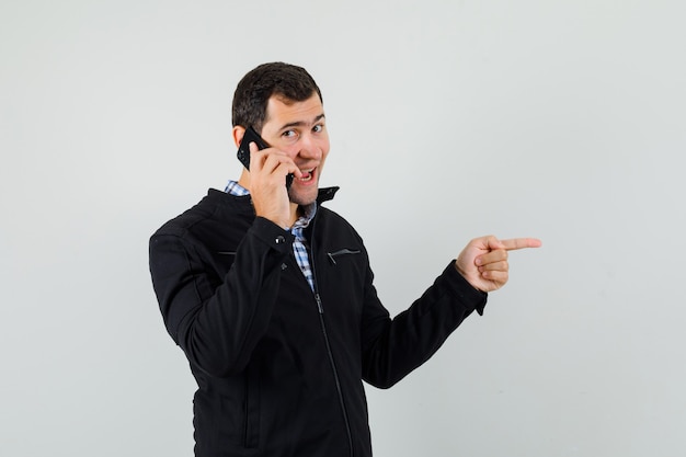 Young man talking on mobile phone, pointing aside in shirt, jacket and looking happy. front view.