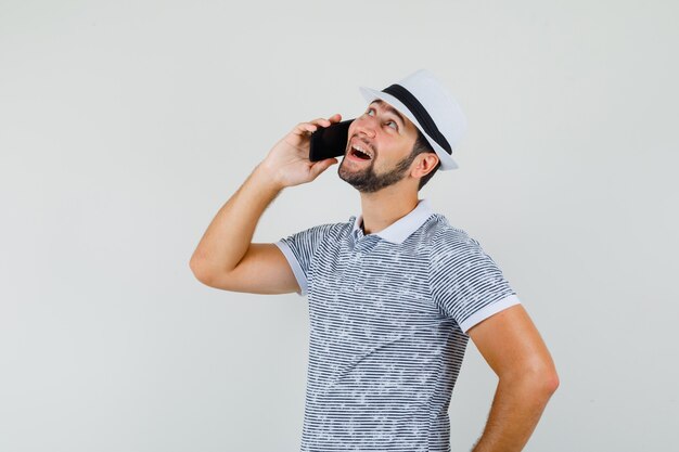 Young man talking on mobile phone in t-shirt, hat and looking cheerful. front view.
