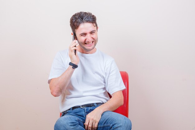 T- 셔츠, 청바지에 의자에 앉아 기뻐 보이는 동안 휴대 전화로 얘기하는 젊은 남자