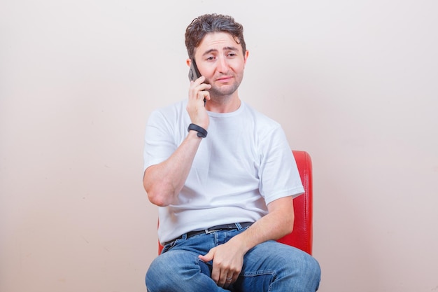 Young man talking on mobile phone while sitting on chair in t-shirt, jeans