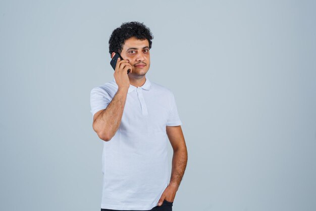 Young man talking on mobile phone in white t-shirt and looking hopeful , front view.