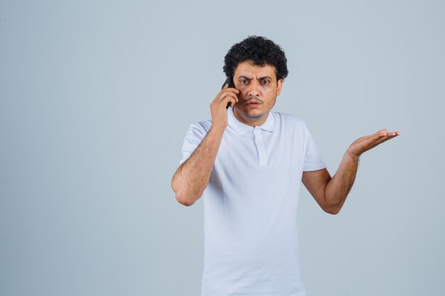 Молодой человек разговаривает по мобильному телефону в белой футболке и недоумевает, вид спереди.