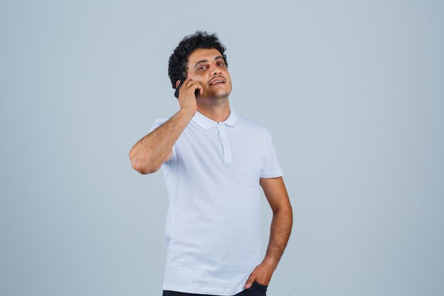 Молодой человек разговаривает по мобильному телефону в белой футболке и выглядит гордым. передний план.