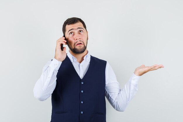 Young man talking on mobile phone with spread palm in shirt and vest and looking helpless