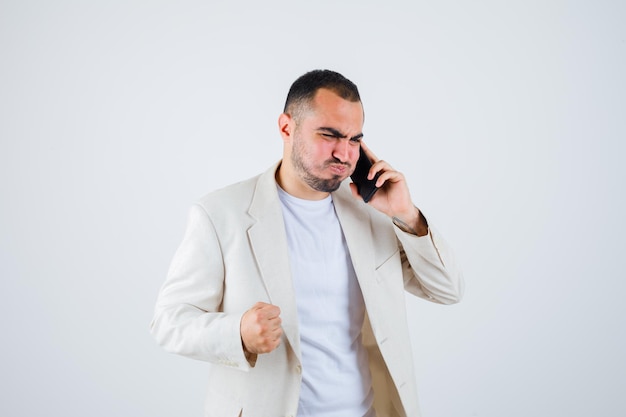Young man talking to phone, clenching fist in white t-shirt, jacket and looking harried. front view.