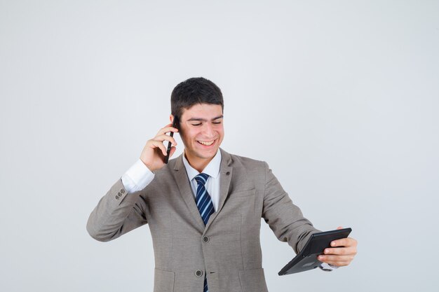 Young man talking to phone, looking at calculator in formal suit