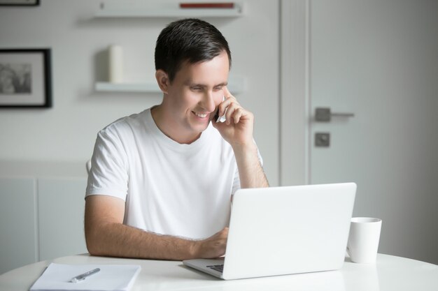 Young man talking on phone at the white desk working at laptop