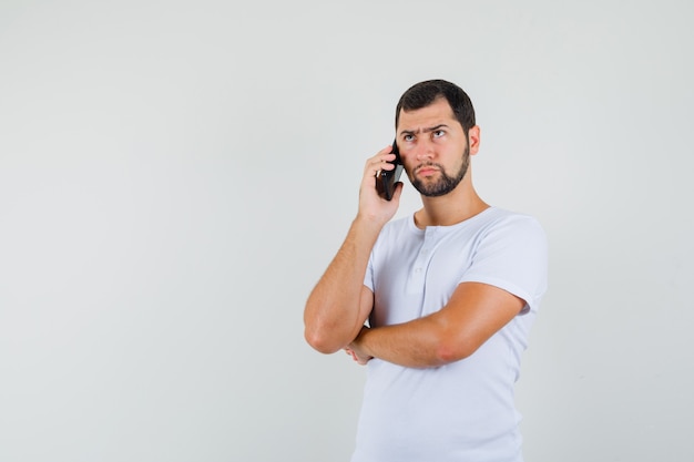 Young man talking on phone in white t-shirt and looking worried , front view. space for text