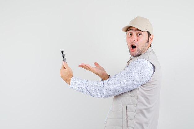 Young man talking to someone via videocall in beige jacket and cap and looking surprised , front view.