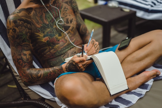a young man in tattoos wearing headphones listens to music and draws in a notebook.