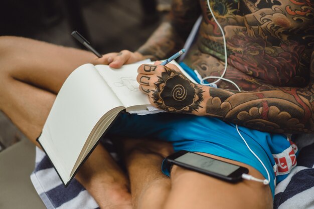 молодой человек в татуировках в наушниках слушает музыку и рисует в блокноте.