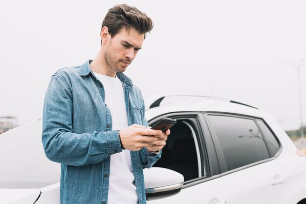 Young man texting message on cellphone standing near the car window