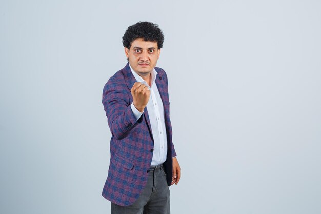 Young man threatening with fist in shirt, jacket, pants and looking powerful , front view.