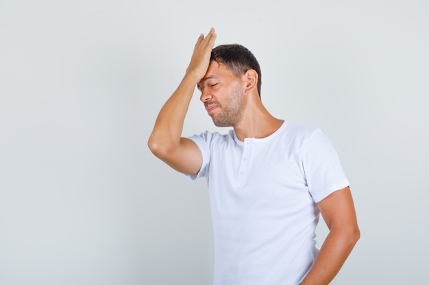 Young man touching forehead with palm in white t-shirt and looking forgetful, front view.