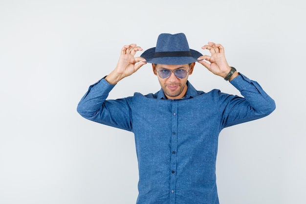 Young man touching his hat in blue shirt and looking elegant. front view.