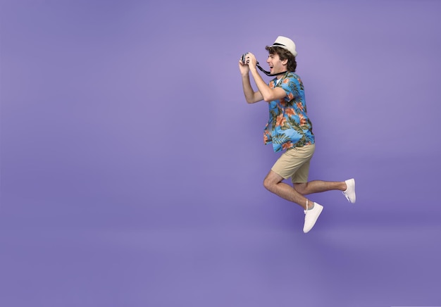 Young man tourist smiling and jumping with camera in hand isolated on purple background