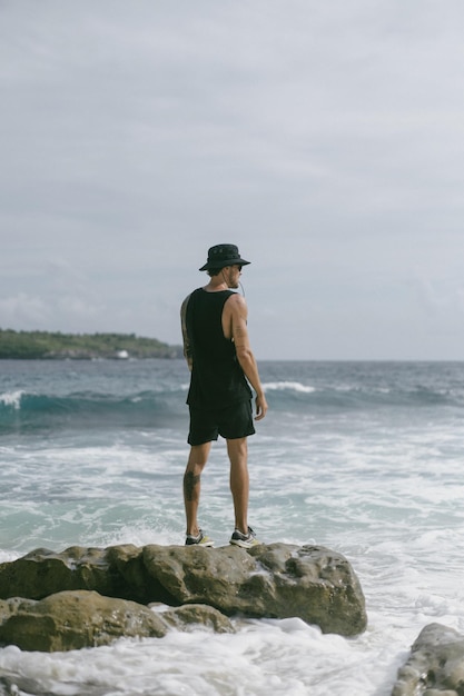 Free photo a young man traveler with a backpack on the ocean on the rocks looks into the distance.