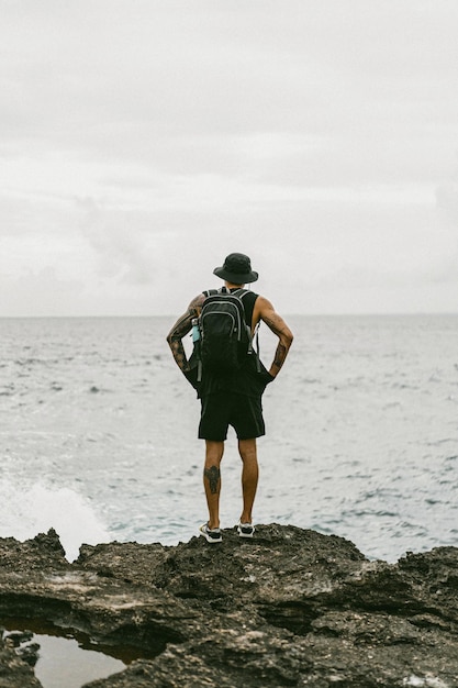 A young man traveler with a backpack on the ocean on the rocks looks into the distance.