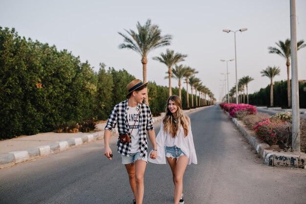 Young man in trendy hat and denim shorts holding girlfriend's hand, walking down the street with bushes on the side. Beautiful couple in stylish outfit spending time outside, enjoys exotic views