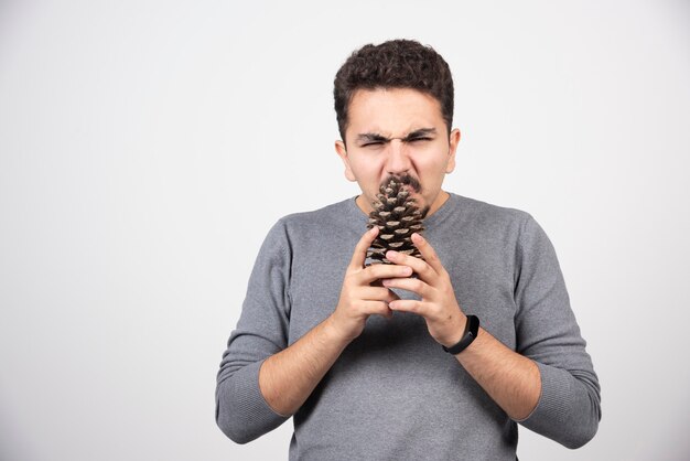 A young man trying to bite pinecone over a white wall.