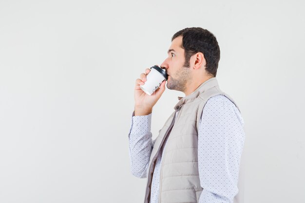 Young man trying to drink takeaway cup of coffee in beige jacket and looking serious , front view.