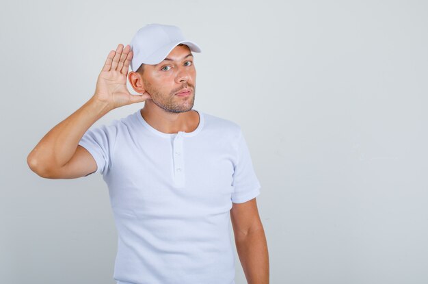 흰색 t- 셔츠, 모자 전면보기에서 귀 제스처와 함께 듣고 노력하는 젊은 남자.