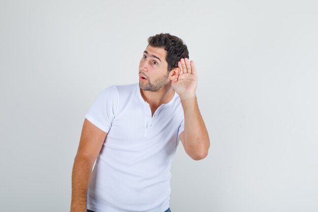 Young man trying to listen something confidential in white t-shirt and looking cunning