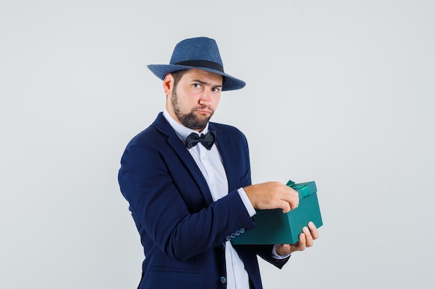 Young man trying to open present box in suit, hat and looking hesitant. front view.