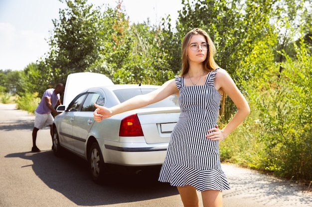 若い女性がヒッチハイクをしている間に車を修理しようとしている若い男