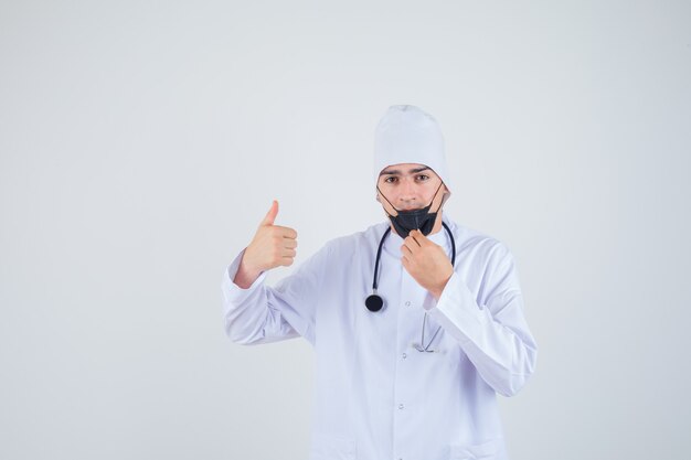 Young man trying yo take off mask, showing thumb up in white uniform, mask and looking satisfied