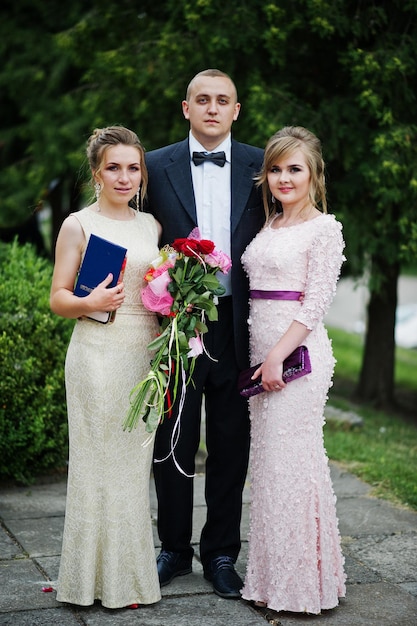 Free photo young man in tuxedo posing with two girls in evening gowns on the graduation ceremony