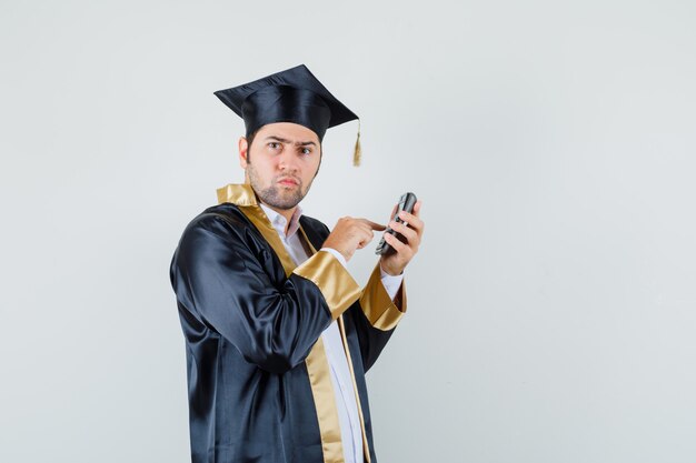 Young man using calculator in graduate uniform and looking pensive , front view.