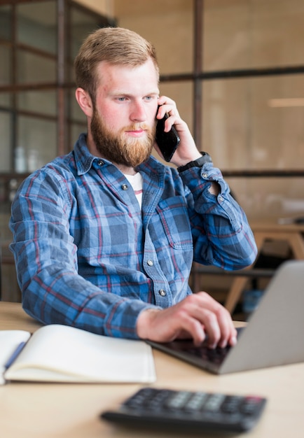 Young man using cellphone while working on laptop in office