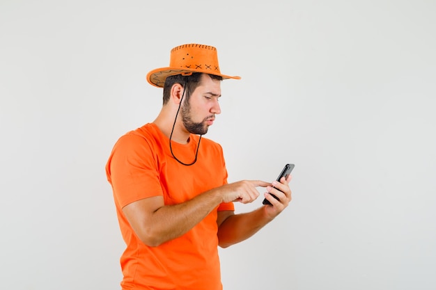 オレンジ色のTシャツ、帽子、忙しそうに見える携帯電話を使用して若い男
