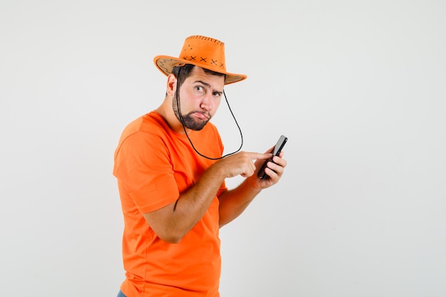 オレンジ色のTシャツ、帽子、物思いにふける携帯電話を使用して若い男。